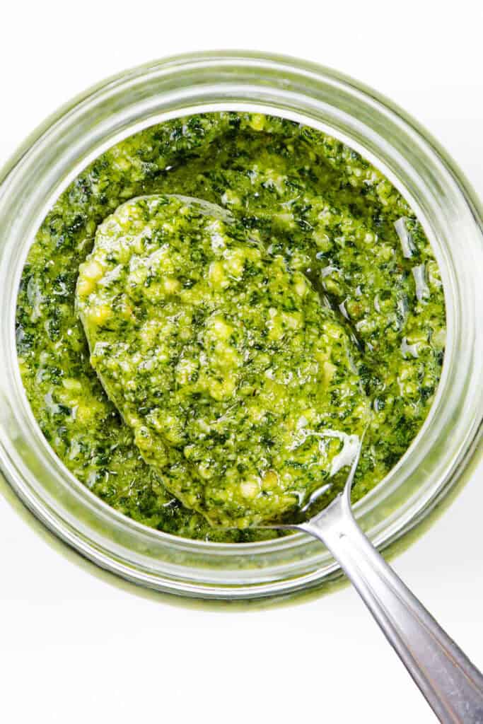 A jar filled with batch pesto sauce, with a silver spoon dipped into it. The vibrant mixture boasts a coarse texture, featuring ingredients like basil, garlic, pine nuts, and olive oil. This quick & easy delight sits on a white surface.