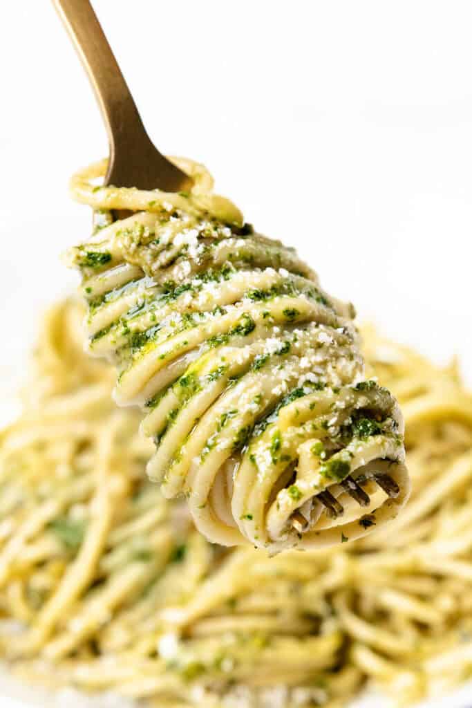 A fork twirls spaghetti coated in vibrant green pesto, quickly sprinkled with grated cheese. In the background, a blurred plate of this quick and easy pasta dish awaits.