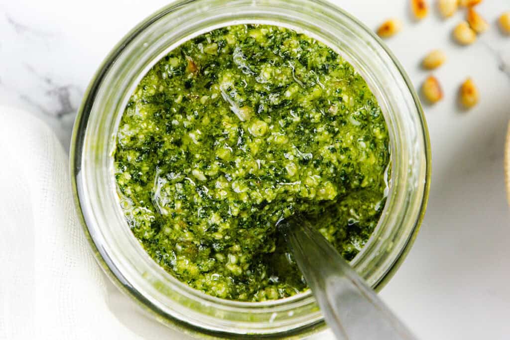 A top view of a jar filled with vibrant, batch pesto sauce. A spoon is dipped into the mixture, showcasing its coarse texture. A few pine nuts are scattered on the white marble surface beside the jar, emphasizing how quick & easy it is to elevate any meal with this delicious pesto.