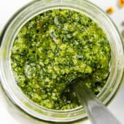 A top view of a jar filled with vibrant, batch pesto sauce. A spoon is dipped into the mixture, showcasing its coarse texture. A few pine nuts are scattered on the white marble surface beside the jar, emphasizing how quick & easy it is to elevate any meal with this delicious pesto.