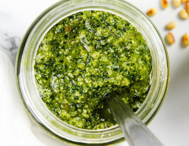 A top view of a jar filled with vibrant, batch pesto sauce. A spoon is dipped into the mixture, showcasing its coarse texture. A few pine nuts are scattered on the white marble surface beside the jar, emphasizing how quick & easy it is to elevate any meal with this delicious pesto.