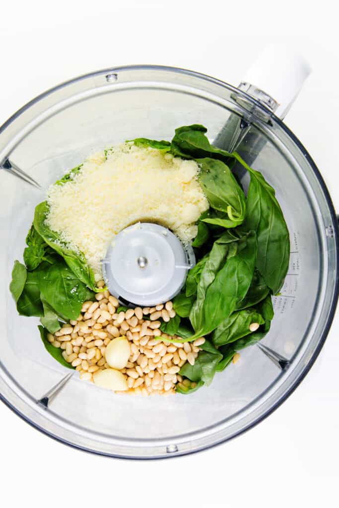 A food processor bowl filled with fresh basil leaves, grated Parmesan cheese, pine nuts, and garlic cloves, ready to be blended for an easy pesto recipe.