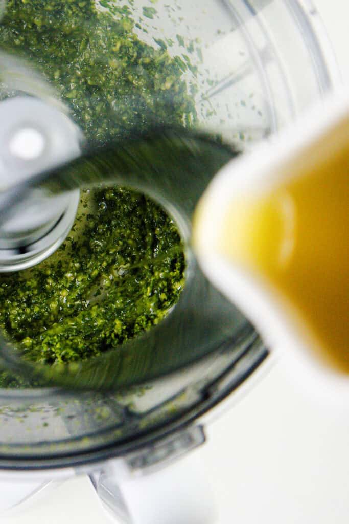 Close-up of a food processor making a quick batch pesto. A stream of olive oil flows into the mixture, visible through the processor's transparent lid.