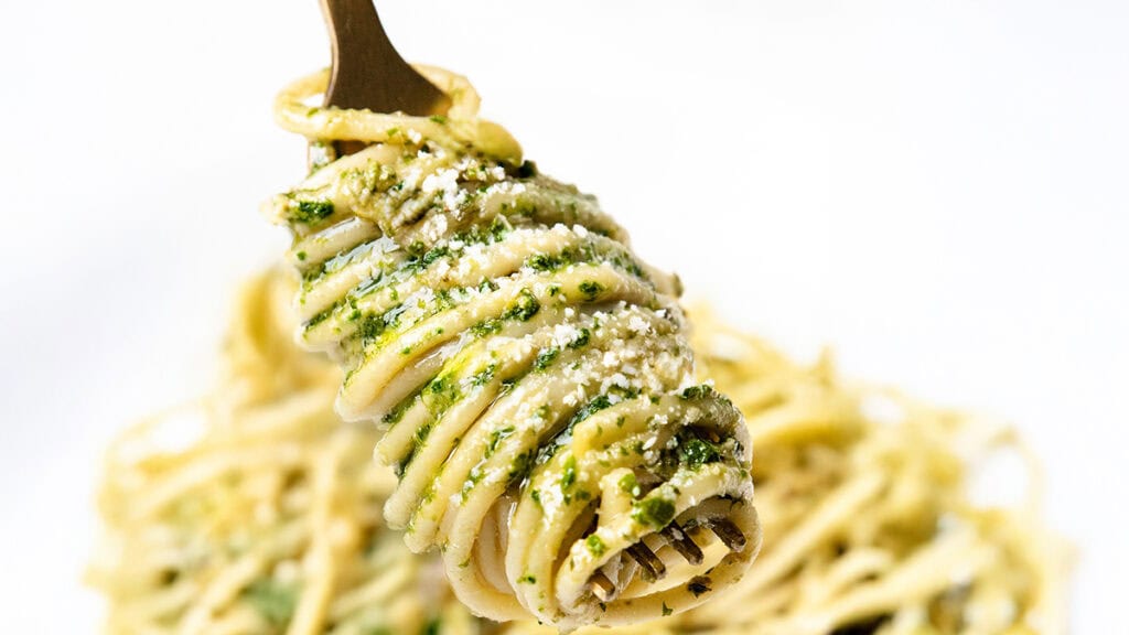 A fork twirls spaghetti with green pesto sauce, garnished with grated cheese, against a white background, showcasing the elegance of restaurant-quality recipes.