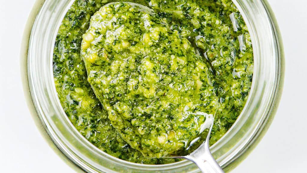 A top view reveals a glass jar brimming with vibrant green pesto, featuring a silver spoon dipped in. This chunky and fresh pasta sauce showcases visible herbs and oil, creating an appetizing look perfect for any dish.