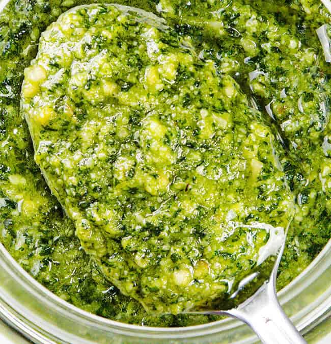 A top view reveals a glass jar brimming with vibrant green pesto, featuring a silver spoon dipped in. This chunky and fresh pasta sauce showcases visible herbs and oil, creating an appetizing look perfect for any dish.