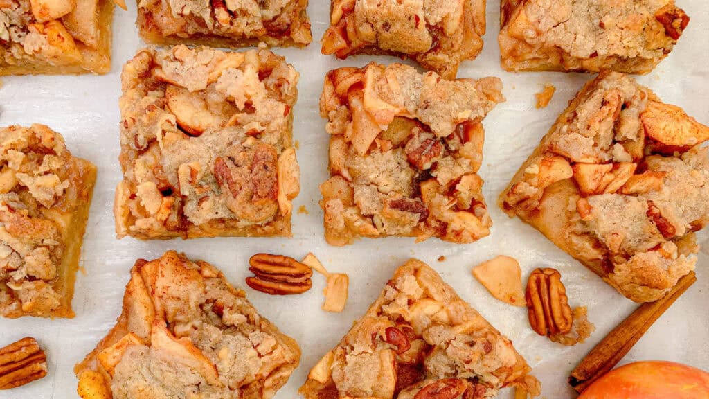 Top view of apple crumble bars with a brown sugar oat topping, arranged in a grid. Slices reveal an apple and cinnamon filling with scattered pecans. A cinnamon stick and apple sit beside, evoking warm Southern cuisine vibes perfect for an autumnal feast.