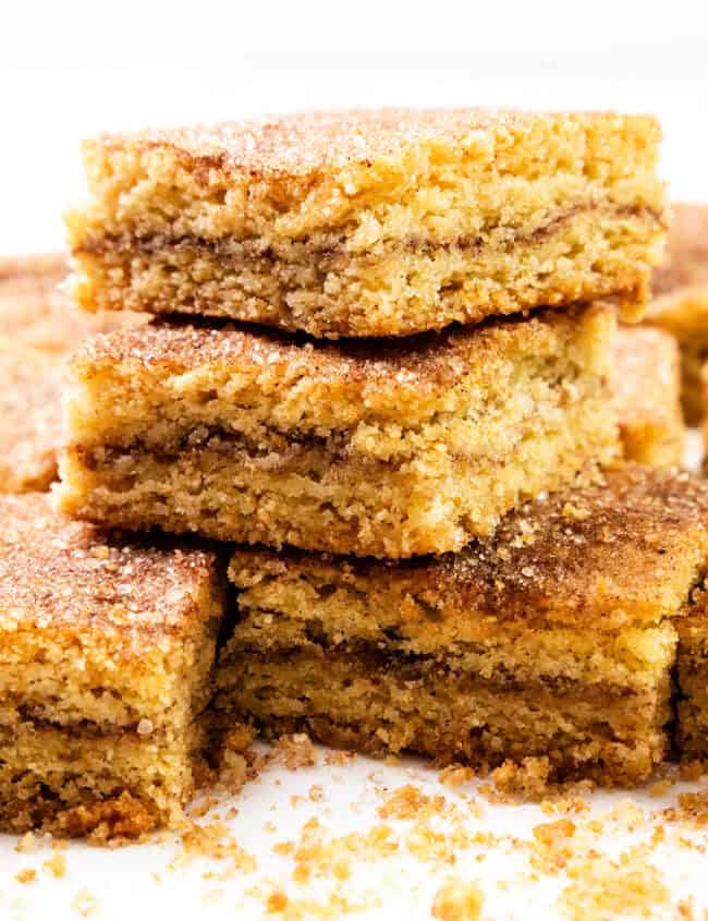 A stack of three Snickerdoodle Bars rests on a white surface, with more bars and crumbs scattered around. These treats boast a golden-brown crust and visibly tempting layers of cinnamon and sugar.