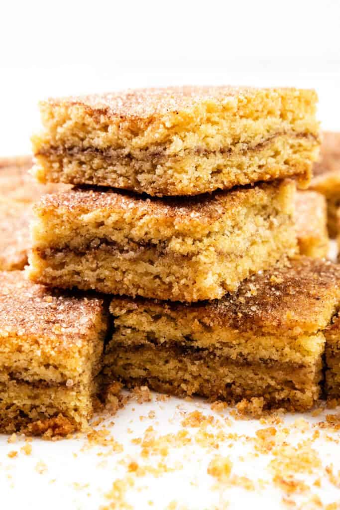 A stack of three Snickerdoodle Bars rests on a white surface, with more bars and crumbs scattered around. These treats boast a golden-brown crust and visibly tempting layers of cinnamon and sugar.