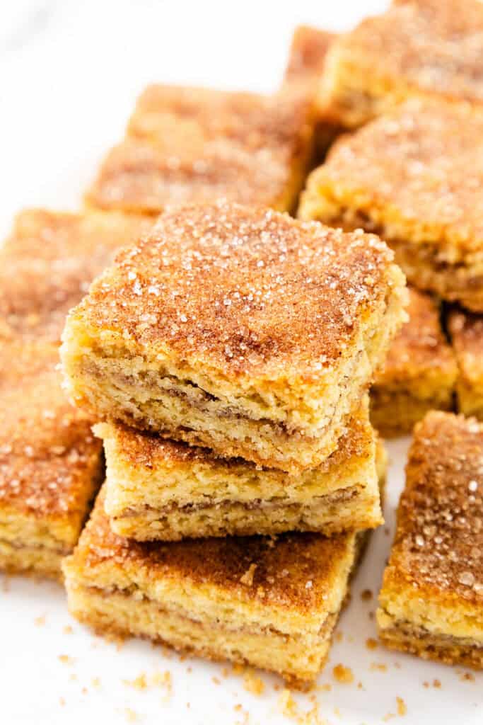 Close-up of a stack of snickerdoodle bars on a white plate. The squares have a golden-brown crust with visible layers, dusted with cinnamon and sugar for a warm, crispy appearance. Some crumbs are scattered around, inviting you to enjoy this delightful dessert bar treat.