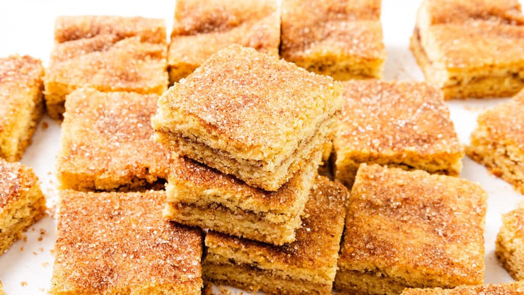 A stack of cinnamon sugar Snickerdoodle Bars takes center stage, surrounded by more delectable pieces. These bar-style treats boast a golden brown finish with a sugary, cinnamon coating on top. Perfectly cut into squares, they belong in any Bar & Brownie Recipes collection.