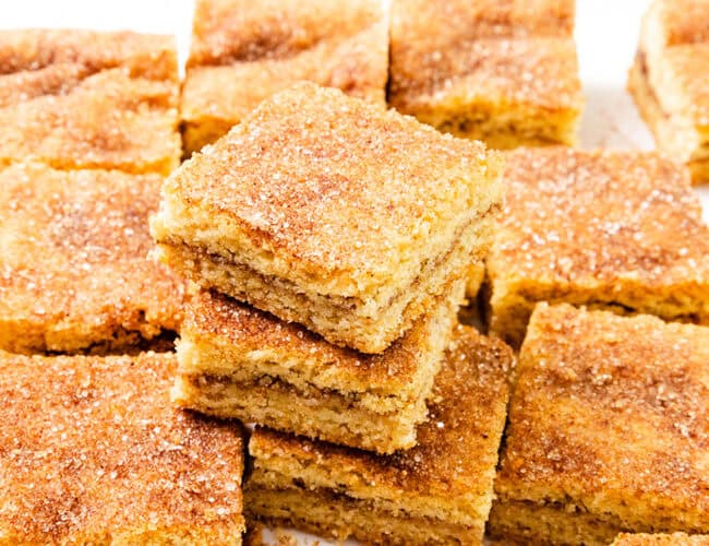 A stack of cinnamon sugar Snickerdoodle Bars takes center stage, surrounded by more delectable pieces. These bar-style treats boast a golden brown finish with a sugary, cinnamon coating on top. Perfectly cut into squares, they belong in any Bar & Brownie Recipes collection.