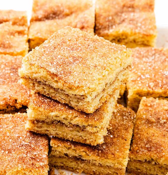 A stack of cinnamon sugar Snickerdoodle Bars takes center stage, surrounded by more delectable pieces. These bar-style treats boast a golden brown finish with a sugary, cinnamon coating on top. Perfectly cut into squares, they belong in any Bar & Brownie Recipes collection.