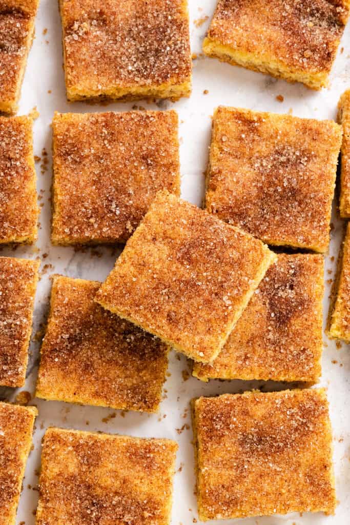 A batch of cinnamon sugar squares, reminiscent of classic Snickerdoodle Bars, is arranged neatly. Each dessert bar has a golden-brown crust, dusted with a layer of cinnamon and sugar, creating a warm, rustic appearance. They're placed on a white surface.