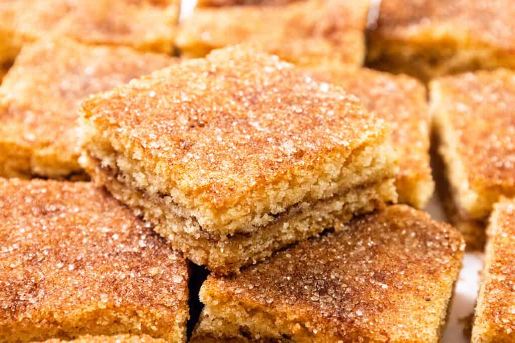 Close-up of several square Snickerdoodle Bars stacked and arranged neatly. The squares boast a golden-brown crust with a visible layer of cinnamon sugar on top, giving them a textured appearance.