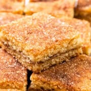 Close-up of several square Snickerdoodle Bars stacked and arranged neatly. The squares boast a golden-brown crust with a visible layer of cinnamon sugar on top, giving them a textured appearance.