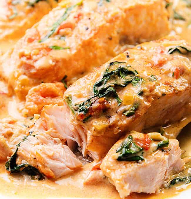 Close-up of creamy Tuscan salmon fillets garnished with spinach and sun-dried tomatoes. A golden fork slices through the flaky, saucy fish, unveiling its tender texture. The dish graces a white plate and is a standout among salmon recipes sure to impress.