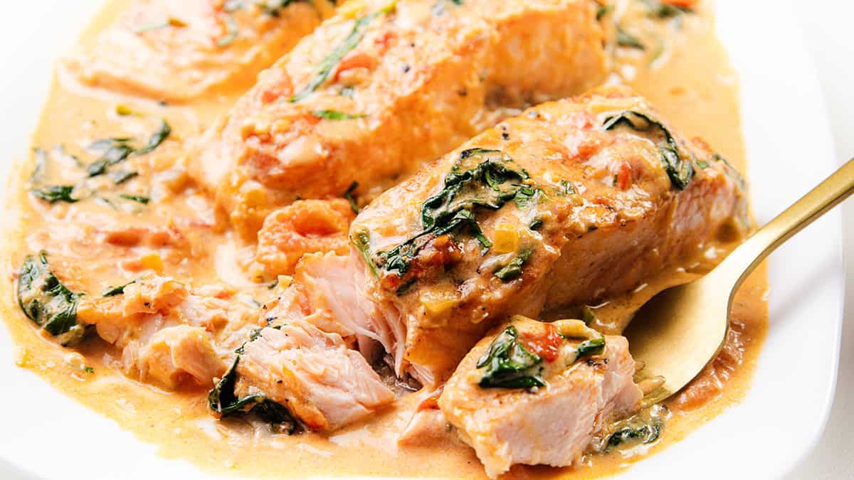 Close-up of creamy Tuscan salmon fillets garnished with spinach and sun-dried tomatoes. A golden fork slices through the flaky, saucy fish, unveiling its tender texture. The dish graces a white plate and is a standout among salmon recipes sure to impress.