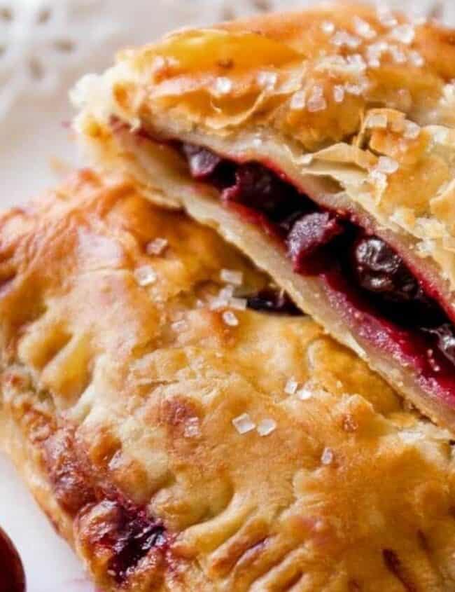 A flaky, golden-brown pastry from the finest cherry recipes is filled with juicy cherries and rests on a decorative white plate. Sugar crystals are sprinkled on top, and a fresh cherry is placed nearby.