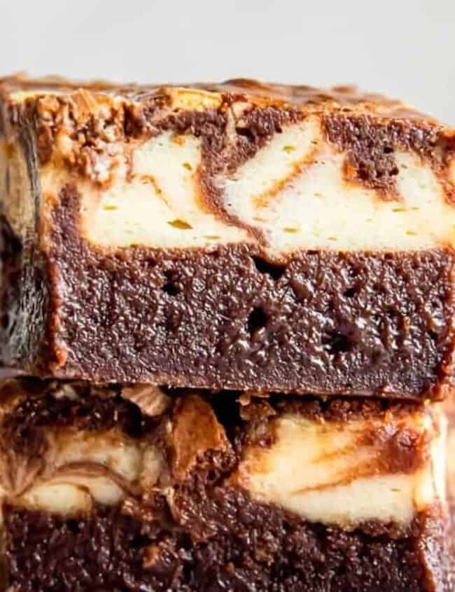 Two stacked marble cheesecake brownies boast a rich, fudgy texture and a creamy, swirled top layer that highlights classic cream cheese indulgence. Other brownies are blurred in the background.