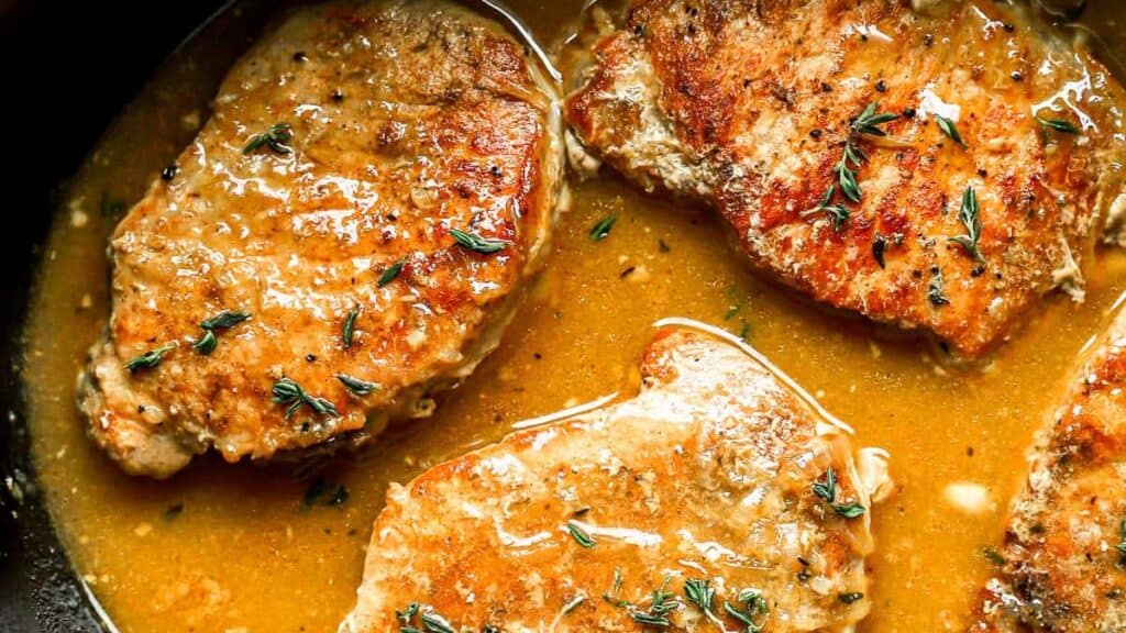 Four golden-brown pork chops are cooked in a rich, savory sauce, garnished with sprigs of fresh thyme. The dish is set in a black pan, showcasing the glossy texture and enticing aroma.