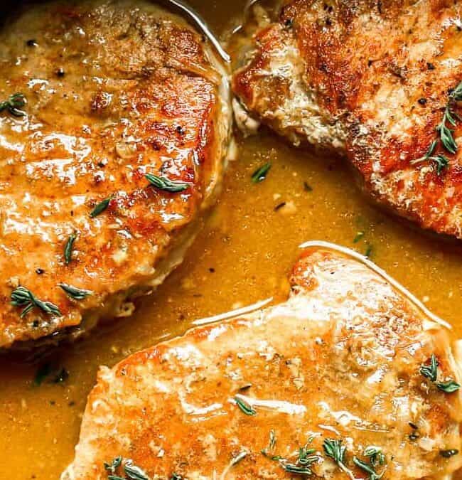 Four golden-brown pork chops are cooked in a rich, savory sauce, garnished with sprigs of fresh thyme. The dish is set in a black pan, showcasing the glossy texture and enticing aroma.