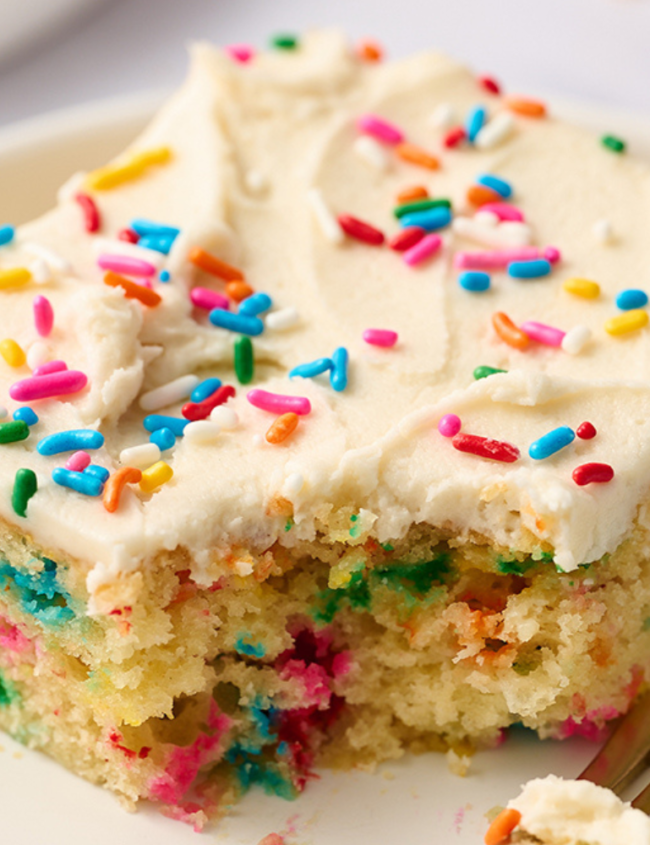 A slice of homemade funfetti cake sits on a white plate with a gold fork. The cake, perfected through beloved cake recipes, is topped with creamy frosting and colorful sprinkles.