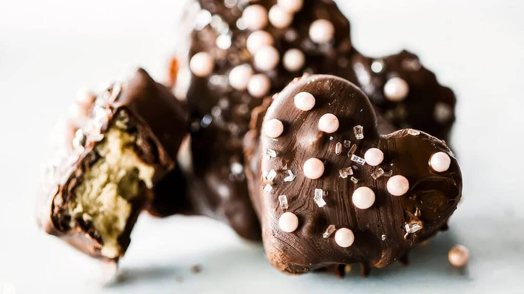 Heart-shaped chocolate candies, perfect for any chocolate recipes, are adorned with pearl-like and sugar crystal sprinkles. A cut piece reveals a creamy filling. The focus is on the delightful textures and decorations against a light background.