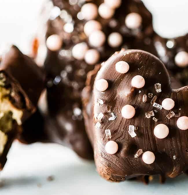 Heart-shaped chocolate candies, perfect for any chocolate recipes, are adorned with pearl-like and sugar crystal sprinkles. A cut piece reveals a creamy filling. The focus is on the delightful textures and decorations against a light background.