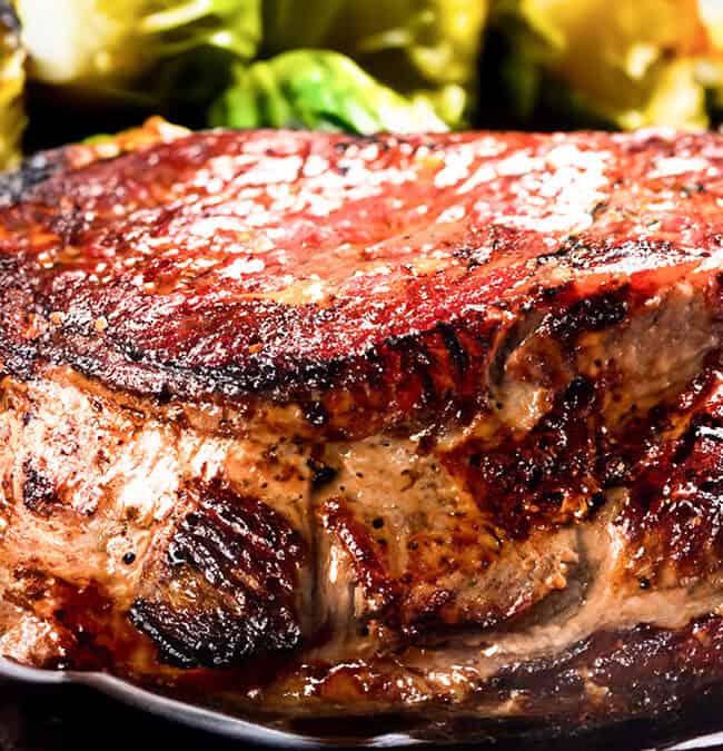 A close-up of a juicy, grilled steak with a crispy, seasoned exterior evokes restaurant-quality recipes. It’s paired with roasted Brussels sprouts in the background, showcasing a rich brown color and a glistening, slightly charred crust.