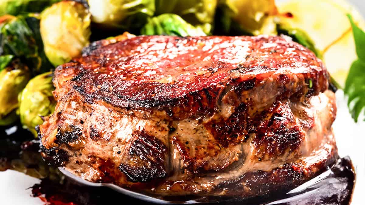 A close-up of a juicy, grilled steak with a crispy, seasoned exterior evokes restaurant-quality recipes. It’s paired with roasted Brussels sprouts in the background, showcasing a rich brown color and a glistening, slightly charred crust.