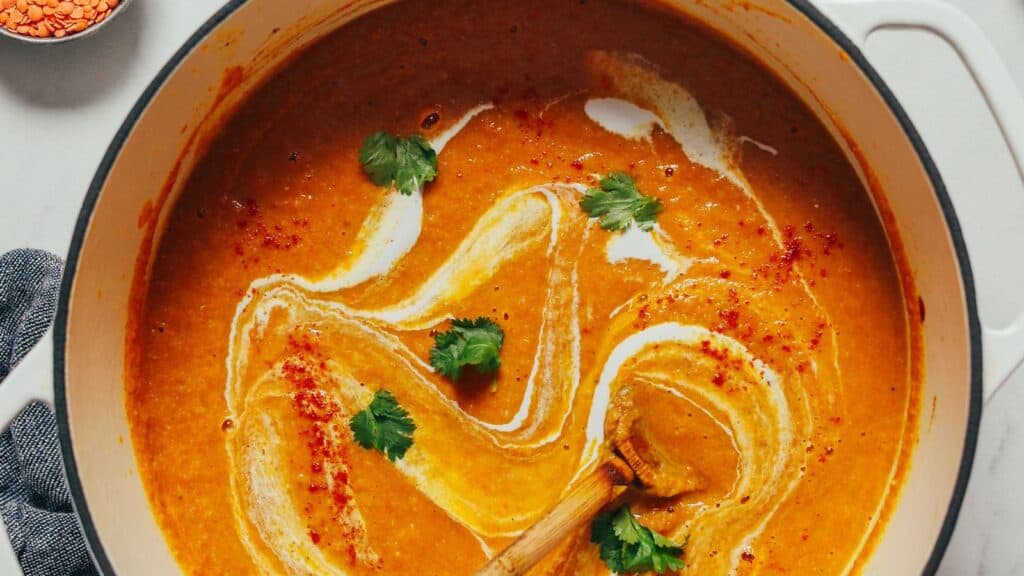 A pot of creamy orange soup garnished with swirls of cream, fresh cilantro leaves, and a sprinkle of red spices, with a wooden spoon resting inside.