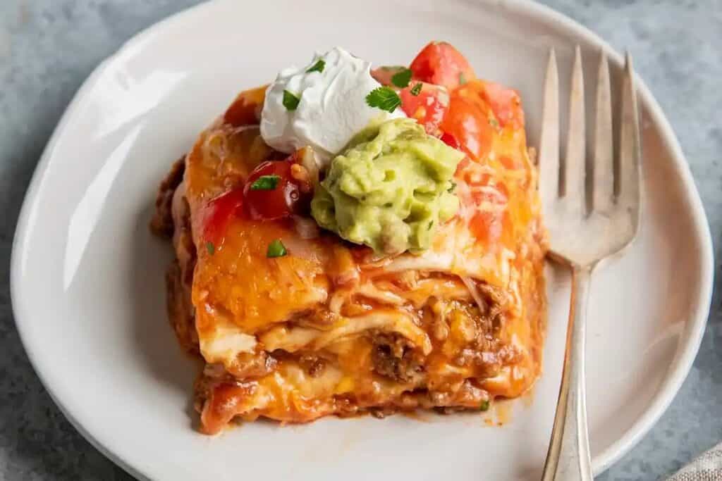 A piece of enchilada casserole featuring ground beef recipes, topped with shredded cheese, a dollop of sour cream, guacamole, diced tomatoes, and cilantro, served on a white plate with a fork.