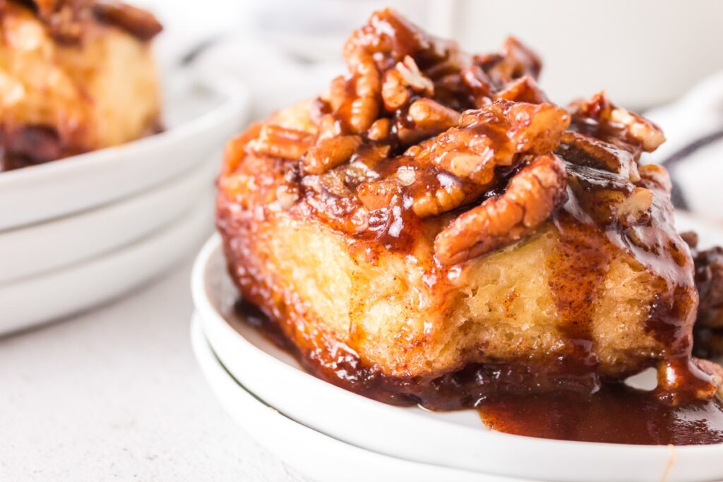 A close-up of a dessert featuring a piece of bread pudding topped with pecans and a rich, caramel sauce, served on a white plate. The glossy, thick sauce enhances the dish's texture and makes every bite worth savoring.