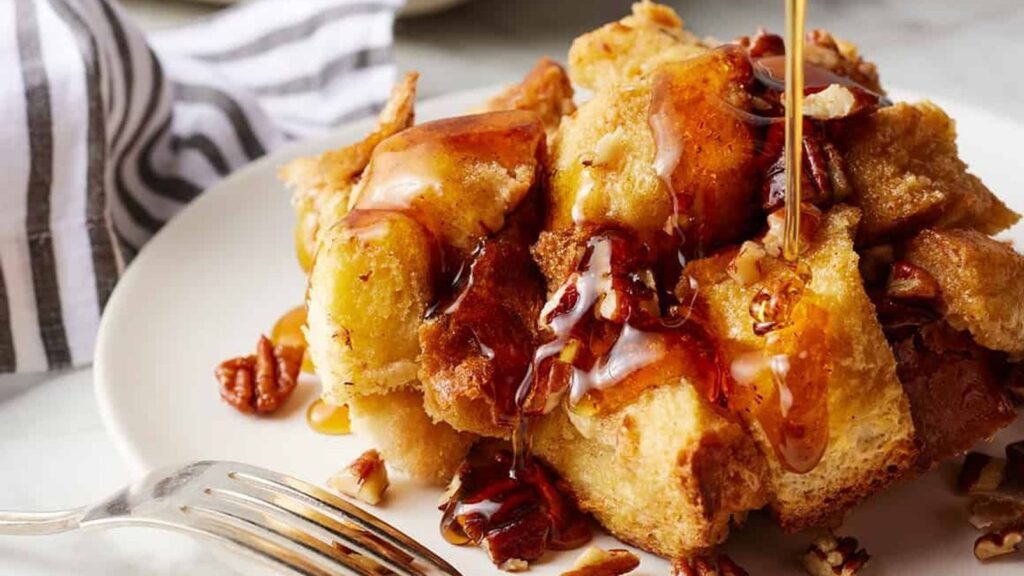 A close-up of a plate of golden-brown French toast cubes, a delightful addition to any brunch recipe collection, topped with pecans and drizzled with maple syrup. A fork rests beside the plate, and a striped napkin is in the background.