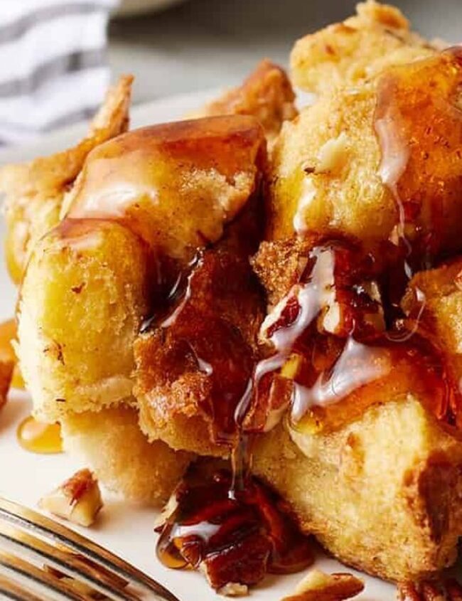 A close-up of a plate of golden-brown French toast cubes, a delightful addition to any brunch recipe collection, topped with pecans and drizzled with maple syrup. A fork rests beside the plate, and a striped napkin is in the background.