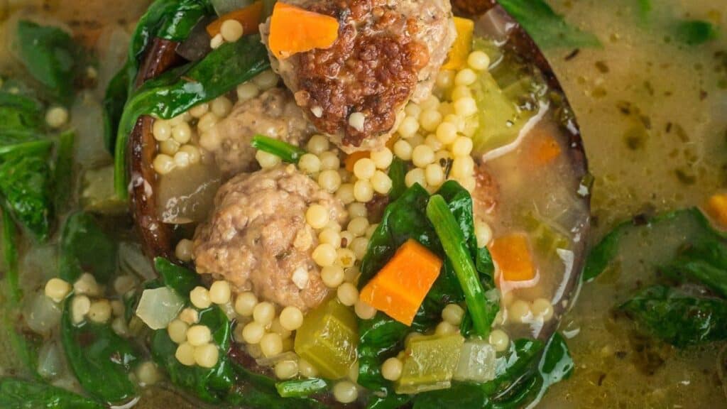 A close-up of a hearty soup recipe featuring meatballs, couscous, spinach, and diced carrots in a rich broth. The ingredients are vibrant and colorful, giving a homemade, comforting appearance.