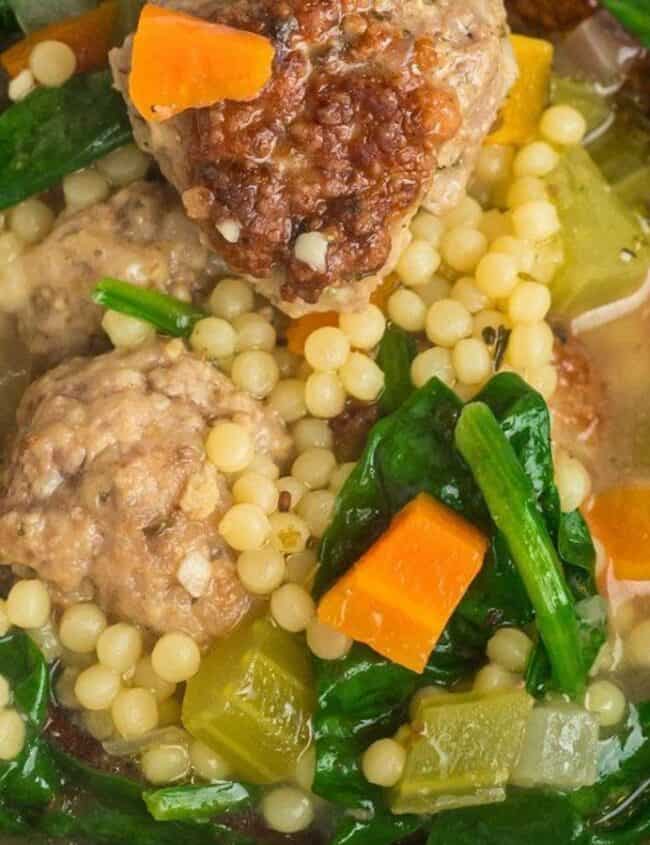 A close-up of a hearty soup recipe featuring meatballs, couscous, spinach, and diced carrots in a rich broth. The ingredients are vibrant and colorful, giving a homemade, comforting appearance.