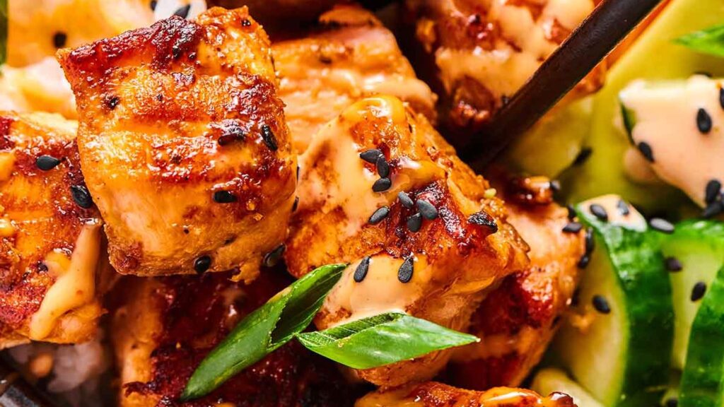 Close-up of grilled chicken pieces topped with a creamy sauce and black sesame seeds. Slices of cucumber and green onions add color and texture, reminiscent of vibrant salmon recipes that celebrate freshness.