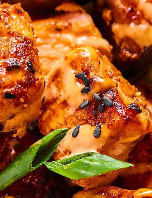 Close-up of grilled chicken pieces topped with a creamy sauce and black sesame seeds. Slices of cucumber and green onions add color and texture, reminiscent of vibrant salmon recipes that celebrate freshness.