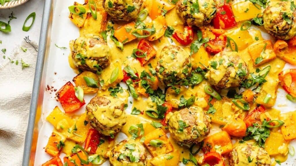 Enjoy these baked chicken meatballs on a sheet pan with colorful bell peppers, topped with a creamy yellow sauce, chopped green onions, and fresh herbs for an easy sheet pan recipe.