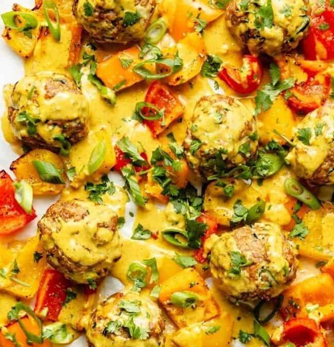 Enjoy these baked chicken meatballs on a sheet pan with colorful bell peppers, topped with a creamy yellow sauce, chopped green onions, and fresh herbs for an easy sheet pan recipe.