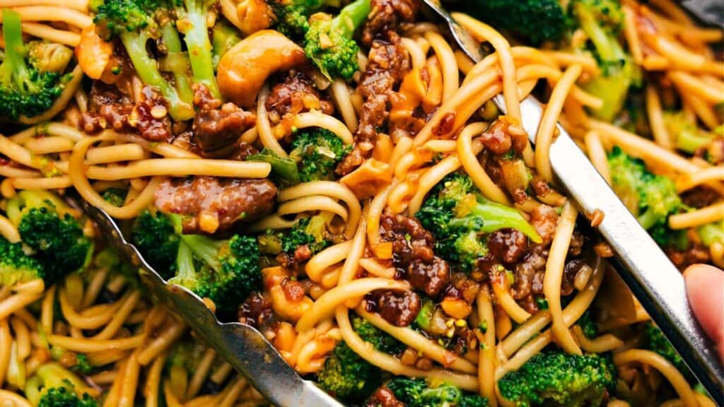 Close-up of a vibrant stir-fry, an effortless eat featuring lo mein noodles, savory beef chunks, and fresh broccoli. Served with metal tongs, this dish highlights an appetizing mix of textures and colors that make dining both simple and delightful.