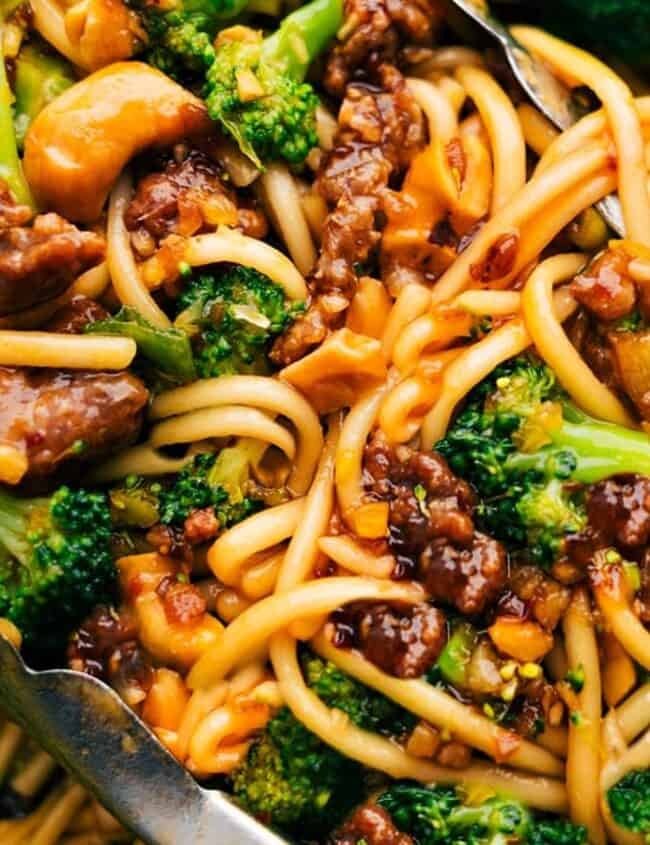 Close-up of a vibrant stir-fry, an effortless eat featuring lo mein noodles, savory beef chunks, and fresh broccoli. Served with metal tongs, this dish highlights an appetizing mix of textures and colors that make dining both simple and delightful.