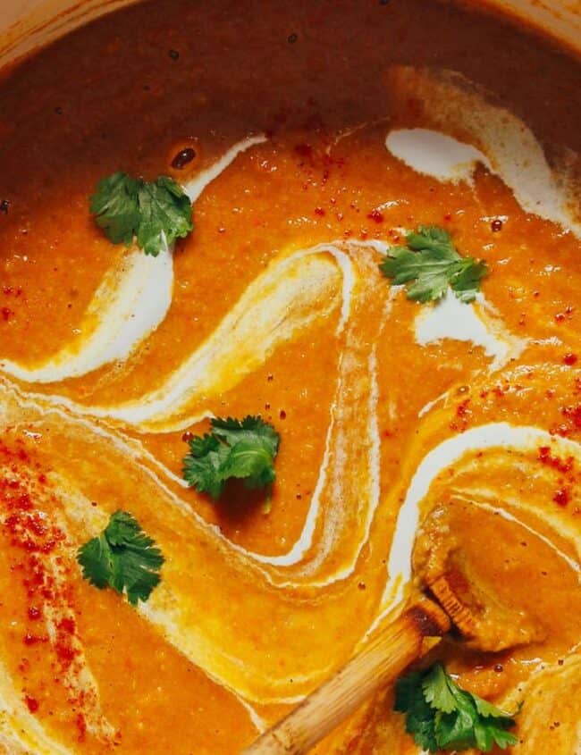 A pot of creamy orange soup garnished with swirls of cream, fresh cilantro leaves, and a sprinkle of red spices, with a wooden spoon resting inside.