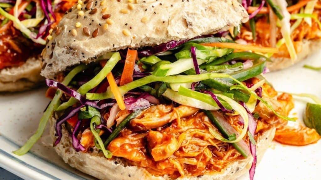 This delightful sandwich features shredded BBQ chicken, cooked to perfection in a slow cooker, and topped with a colorful slaw of green and purple cabbage, carrots, and cucumbers. All served on a seeded bun, the vibrant toppings bring a fresh, crunchy texture to the dish.