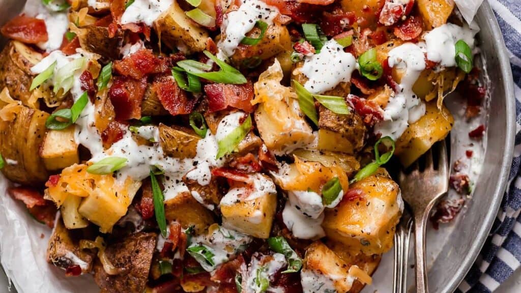 Roasted potatoes topped with crispy bacon, melted cheese, diced green onions, and drizzled with creamy dressing make an irresistible side dish on a metal plate. A fork is placed to the side.