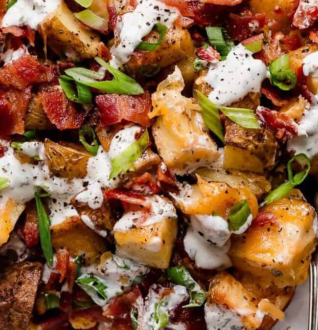 Roasted potatoes topped with crispy bacon, melted cheese, diced green onions, and drizzled with creamy dressing make an irresistible side dish on a metal plate. A fork is placed to the side.