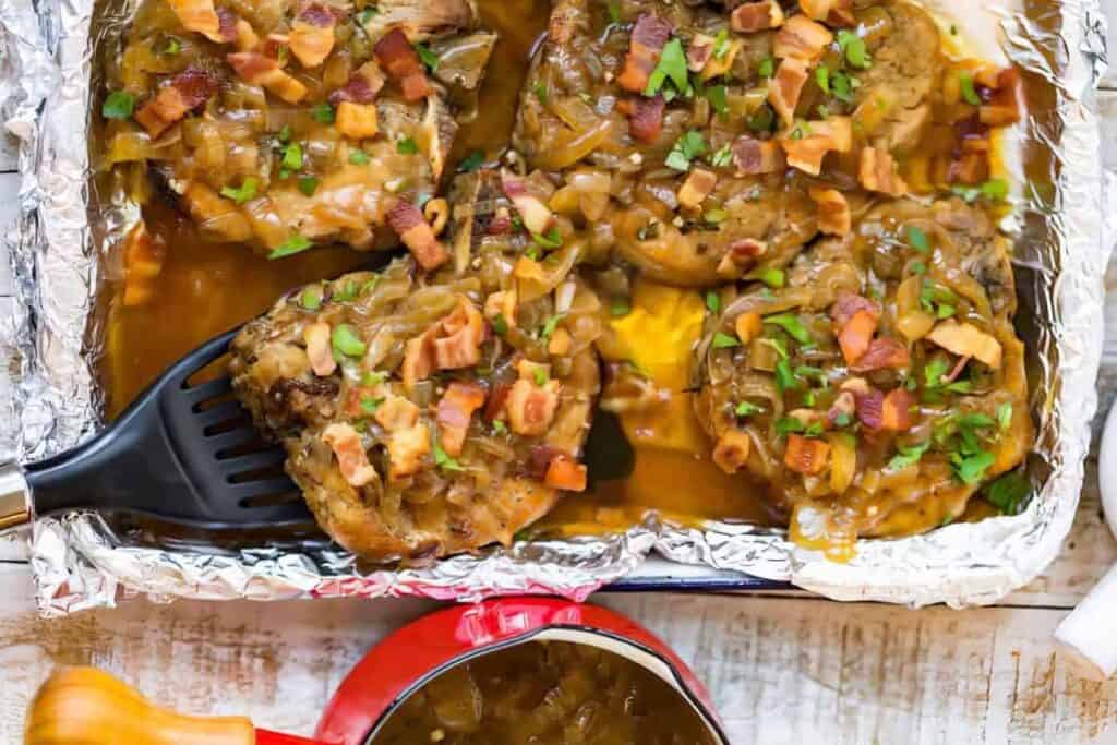 Four baked pork chops with onion gravy, topped with bacon bits and fresh parsley, served in a foil-lined baking dish. A black spatula lifts one chop, showcasing pure comfort food bliss. Nearby, a small red pot holds extra gravy to perfect this delectable pork recipe on a rustic white table.