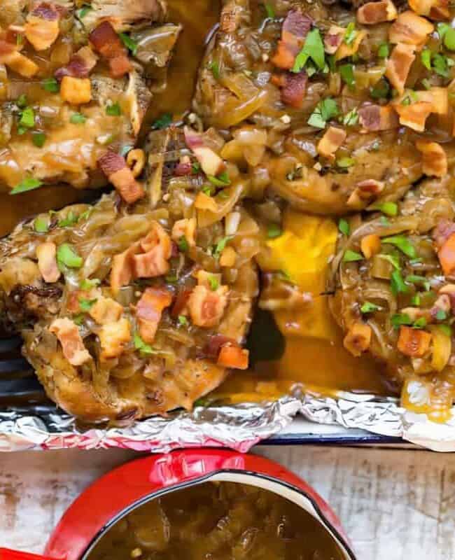 Four baked pork chops with onion gravy, topped with bacon bits and fresh parsley, served in a foil-lined baking dish. A black spatula lifts one chop, showcasing pure comfort food bliss. Nearby, a small red pot holds extra gravy to perfect this delectable pork recipe on a rustic white table.